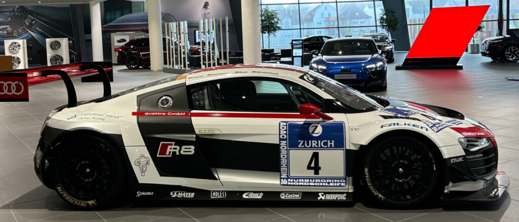 Seitenansicht eines weiß-schwarz-roten Audi R8 Rennwagens mit der Startnummer 4 im Audi Forum Neckarsulm. Das Fahrzeug ist mit Sponsorenlogos von Zurich, Michelin und Akrapovič versehen und besitzt einen markanten großen Heckflügel. Im Hintergrund ist die moderne, verglaste Ausstellungshalle mit weiteren Fahrzeugen zu sehen.