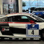 Seitenansicht eines weiß-schwarz-roten Audi R8 Rennwagens mit der Startnummer 4 im Audi Forum Neckarsulm. Das Fahrzeug ist mit Sponsorenlogos von Zurich, Michelin und Akrapovič versehen und besitzt einen markanten großen Heckflügel. Im Hintergrund ist die moderne, verglaste Ausstellungshalle mit weiteren Fahrzeugen zu sehen.