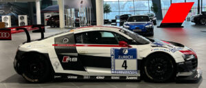Seitenansicht eines weiß-schwarz-roten Audi R8 Rennwagens mit der Startnummer 4 im Audi Forum Neckarsulm. Das Fahrzeug ist mit Sponsorenlogos von Zurich, Michelin und Akrapovič versehen und besitzt einen markanten großen Heckflügel. Im Hintergrund ist die moderne, verglaste Ausstellungshalle mit weiteren Fahrzeugen zu sehen.