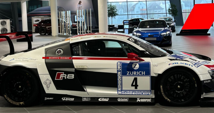 Seitenansicht eines weiß-schwarz-roten Audi R8 Rennwagens mit der Startnummer 4 im Audi Forum Neckarsulm. Das Fahrzeug ist mit Sponsorenlogos von Zurich, Michelin und Akrapovič versehen und besitzt einen markanten großen Heckflügel. Im Hintergrund ist die moderne, verglaste Ausstellungshalle mit weiteren Fahrzeugen zu sehen.