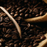 Zu sehen sind Kaffeebohnen, die aus einem Jutesack kommen. Ein Teil der Bohnen liegt in einer Holzkelle.