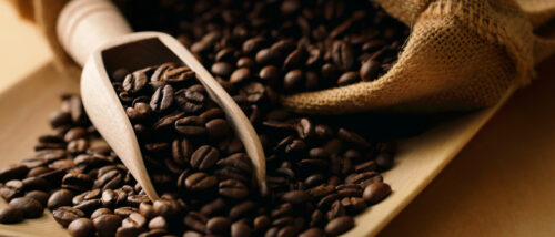 Zu sehen sind Kaffeebohnen, die aus einem Jutesack kommen. Ein Teil der Bohnen liegt in einer Holzkelle.