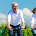 Eine Dame und ein Herr im Rentenalter fahren gemeinsam fröhlich Fahrrad auf einem Weg vor einem grünen Weinberg unter strahlend blauem Himmel.