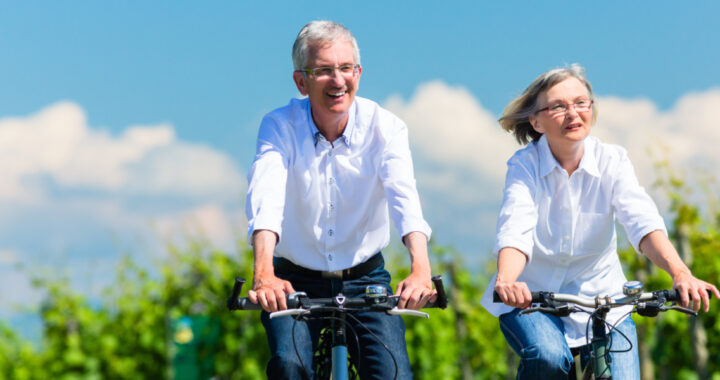 Eine Dame und ein Herr im Rentenalter fahren gemeinsam fröhlich Fahrrad auf einem Weg vor einem grünen Weinberg unter strahlend blauem Himmel.