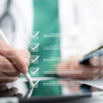 Eine digitale Grafik einer Checkliste mit abgehakten Kästchen schwebt über einem Tablet im Vordergrund. Ein Arzt im unscharfen Hintergrund, bekleidet mit weißem Kittel, grünem Kasack und Stethoskop, bearbeitet die Liste mit einem digitalen Eingabestift.