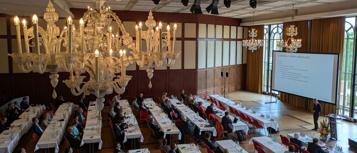 Blick von oben auf Tagung mit Person, die Rede hält und zahlreichen Personen, die zuhören